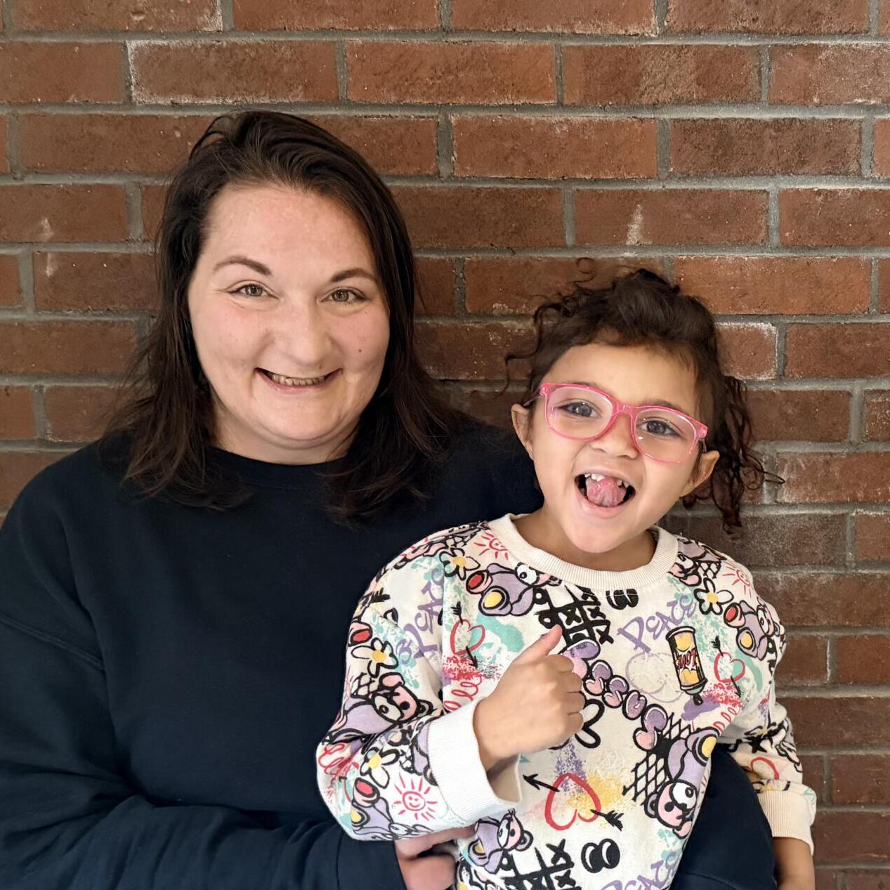 An adult and a young girl with glasses sit together, smiling, in front of a brick wall.