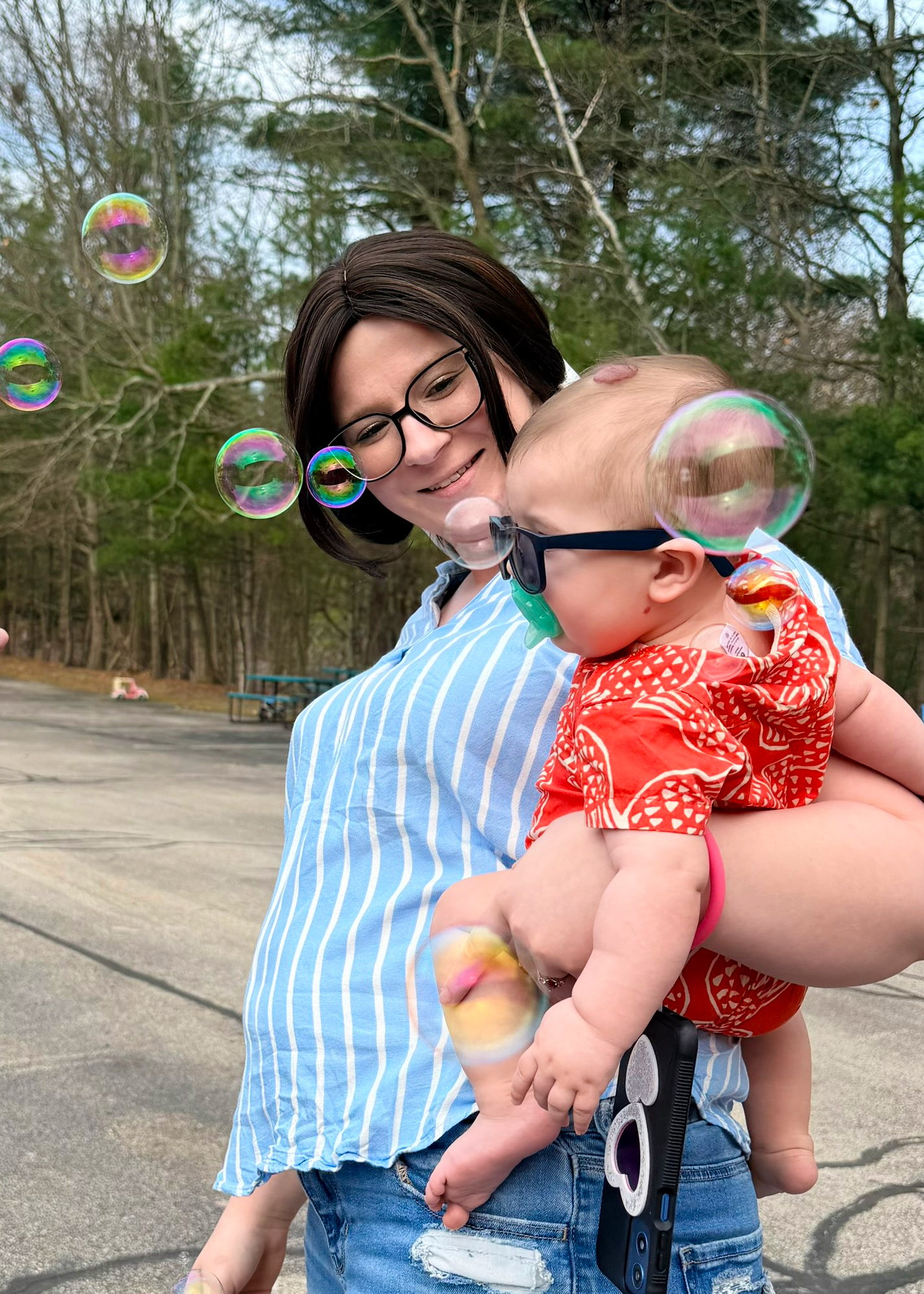 A woman holds a baby dressed in red while standing outdoors. Both are looking at floating soap bubbles. Trees and pavement are in the background.