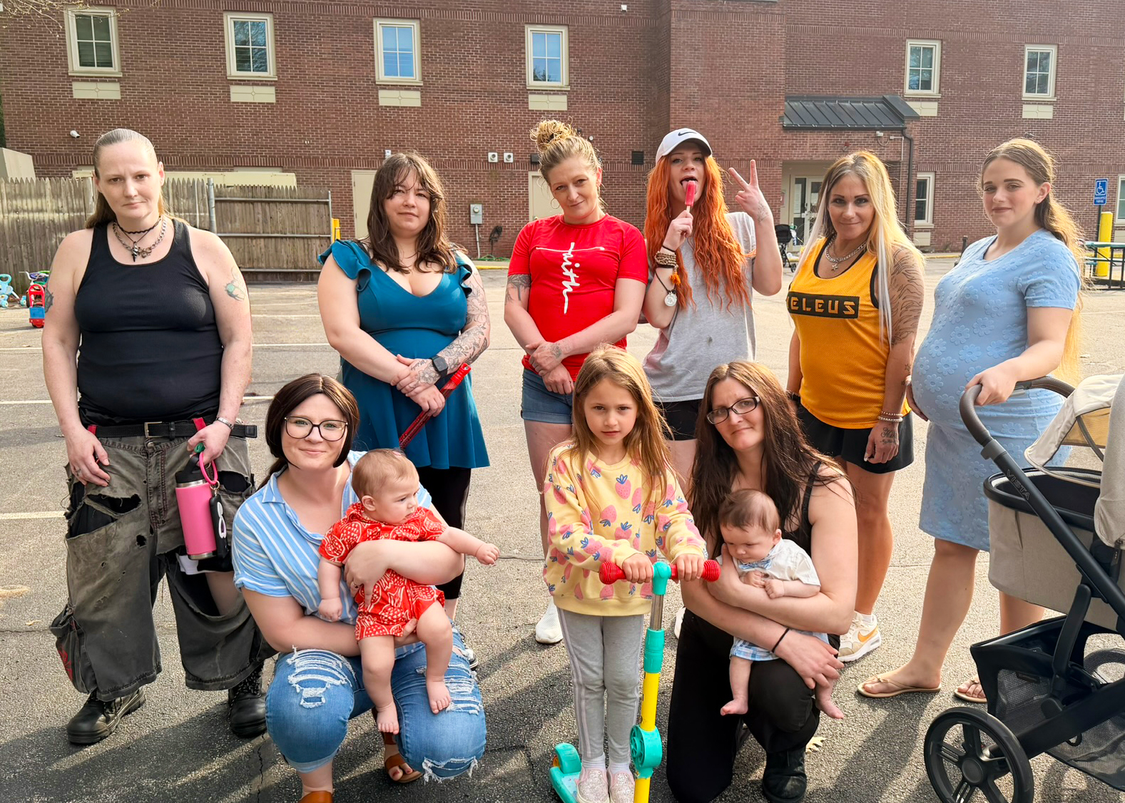 A group of women and children pose outdoors in a parking lot, with some holding babies and one woman pushing a stroller. A brick building is in the background.