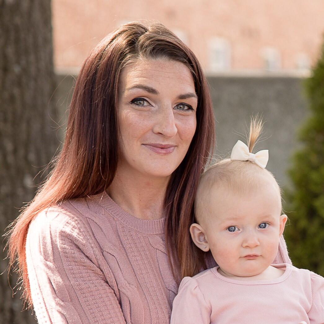 Woman with long brown hair holds a baby with a small ponytail and a bow, both wearing light pink sweaters, sitting outdoors on a bench.